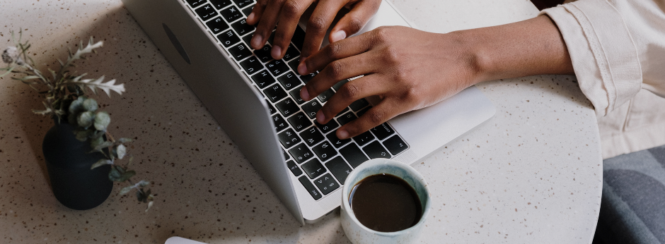 looking down at a white table top with hands typing on a laptop keyboard, a cup of coffee to the side and a small greenery bouquet in a vase behind the laptop