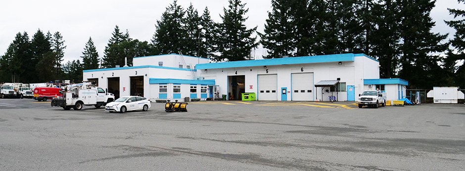 A large paved area with a white mechanic building with blue trim and cars and trucks parked outside of it.