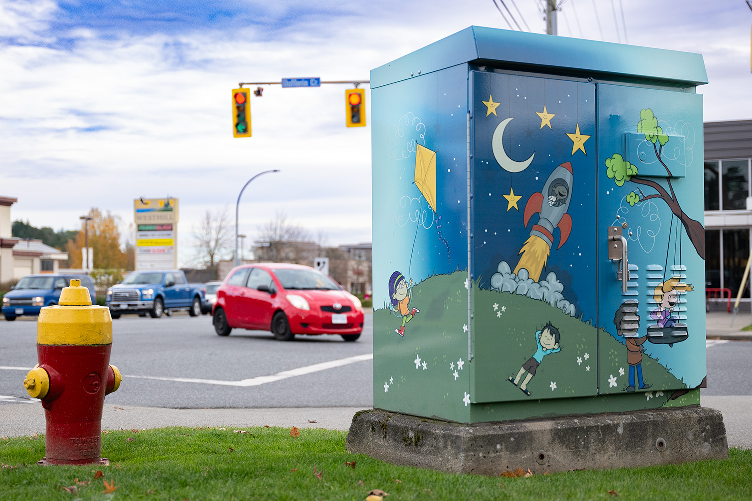 A traffic control cabinet with playful art shows children at imaginative play, flying a kit and even taking off in a rocket ship beneath the starts and crescent moon.